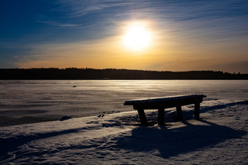 Fototapeta na wymiar Winter landscape picture in Finland. Empty park bench in front. A frozen lake in the background.