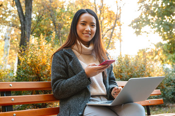 Smiling young asian woman wearing coat sitting on a bench