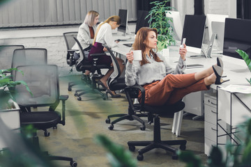 beautiful young businesswoman holding cup and using smartphone while sitting with legs on desk in open space office