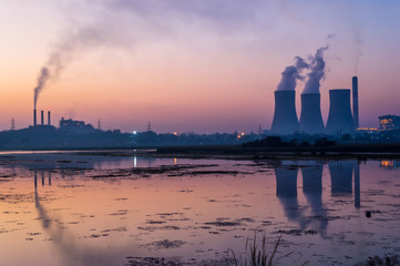 Cooling tower and smoke chimney emitting steam and smoke. Lake near the thermal power plant reflecting.