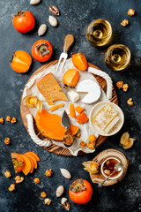 Cheese variety board or platter with cheese assortment, persimmons, honey and nuts. Black stone background. Top view, flat lay
