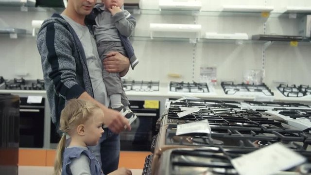 Father holding baby son on arms while cute little daughter opening oven and looking into it in home appliance store