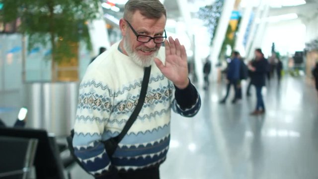Mature man with a gray beard in a knitted sweater and glasses at the airport or train station. The pensioner waves goodbye and leaves carrying a suitcase on wheels behind him