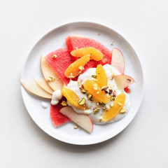 A plate of fresh fruit salad with yogurt, watermelon, orange, apple, and pistachio on a white background, top view