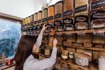 Shopkeeper working in zero waste shop. Shop assistant filling glass jar with red lentils in...