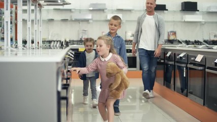 Playful little kids enjoying shopping with father: family walking together in home appliance store and choosing kitchen stove