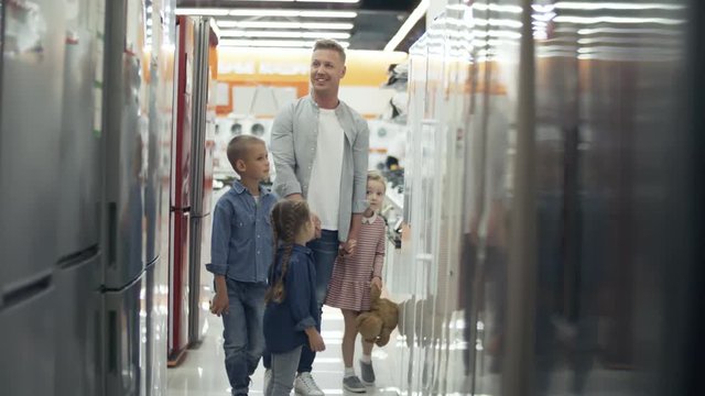 Happy father walking with two little daughters and son along aisle in home appliance store and discussing refrigerators