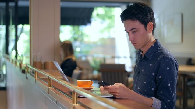 Young Latin American man sitting in coffee shop in Medellin, Colombia, using laptop and smartphone, working or studying online, medium shot. High quality 4K ProRes 422 HQ