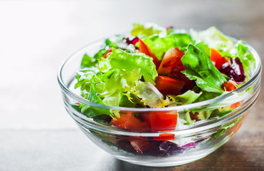 various fresh mix salad leaves with tomato in glass bowl on wooden background.