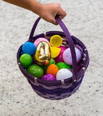 A purple easter egg basket with shiny colored plastic easter eggs in it being held by a child's hand.