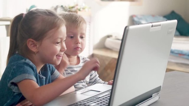 Cute playful little boy and his elder sister smiling, pointing at screen and discussing cartoon on laptop at kitchen table