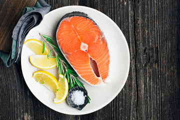 Fresh salmon steak on light plate with lemon and rosemary on wooden background. Top view.