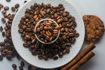 White Coffee cup with roasted coffee beans, cinnamon and biscuits on isolated white background from above.  Coffee beans on plate and in the mug. Cinnamon and cookies on a table with coffee beans.