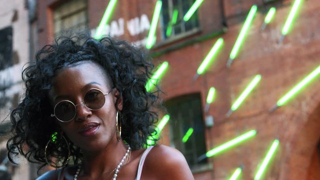 Fashionable young black woman in urban street walking to camera and into focus, close up, low angle, blurred neon strip lights in background