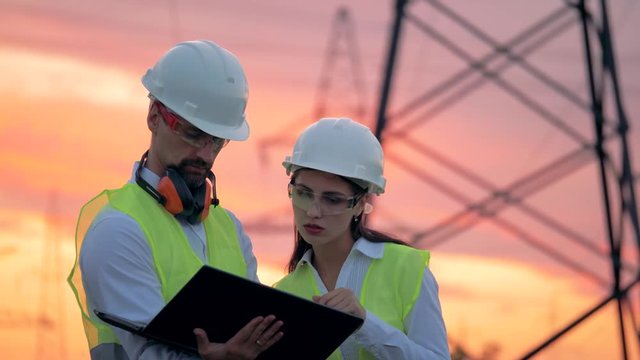 Two engineers are talking in front of electrical transmission lines, working on renewable energy development.