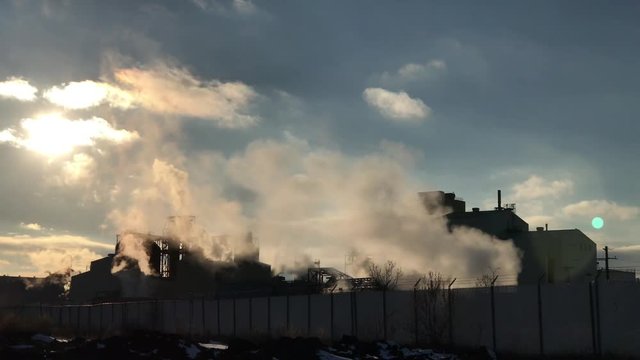 Industrial pollution in winter as hot steam and smoke are emitted into air moving sideways with late afternoon sun hovering above. 