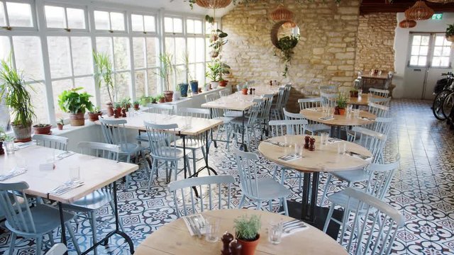 Round tables and duck-egg blue chairs in an empty restaurant with patterned floor tiles and houseplants growing on the window sill
