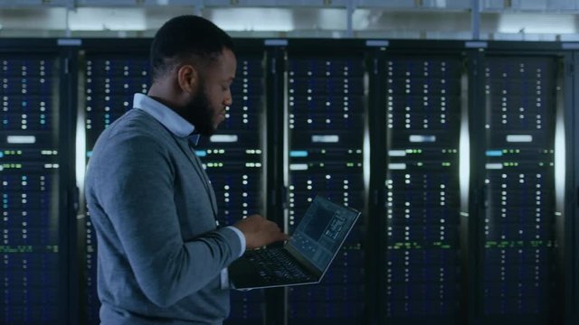 Black Data Center IT Technician Walking Through Server Rack Corridor with a Laptop Computer