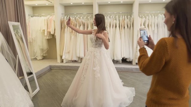 Two brunette girls are in wedding salon. They have a fitting of wedding dress. Both of them are holding phones in hands and making photos.