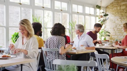 Diners talking and eating at tables in a busy restaurant during the day, handheld