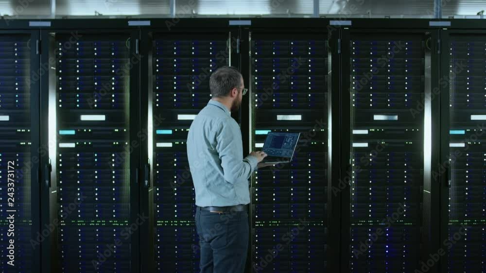 Poster Bearded IT Specialist in Glasses is Working on Laptop in Data Center while Standing Before Server Rack. Running Diagnostics or Doing Maintenance Work. Camera Zooms Out.