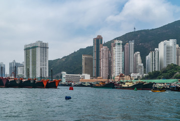 Fototapeta premium Hong Kong, China - May 12, 2010: Wide shot. Plenty black and red fishing vessels docked in harbor with many tall apartment buildings on shore. Clouded sky and green hill in back.