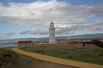 Fototapeta na wymiar Lighthouse on coast of sea, sandy path, in background hills, clouds in sky