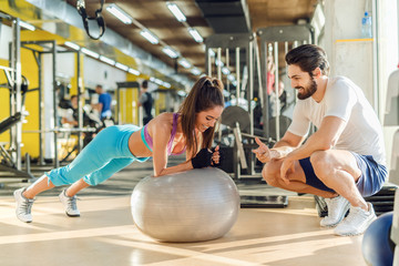 Sporty smiling woman doing planks on pilates ball while her personal trainer crouching next to her and cheering for her.