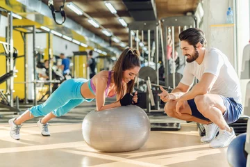 Poster Sporty smiling woman doing planks on pilates ball while her personal trainer crouching next to her and cheering for her. © dusanpetkovic1