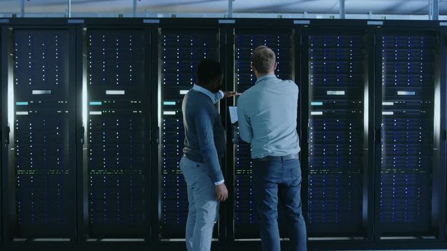 Bearded IT Technician in Glasses with a Laptop Computer and Black Male Engineer Colleague are Talking in Data Center while Working Next to Server Racks. Running Diagnostics or Doing Maintenance Work. 