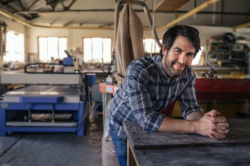 Smiling woodworker leaning on a bench in his large workshop