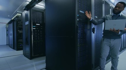 Black IT Technician with a Laptop Computer Gives a Tour to a Young Intern. They Talk in Data Center while Walking Next to Server Racks. Other Colleague is Changing Hard Drives. - Powered by Adobe