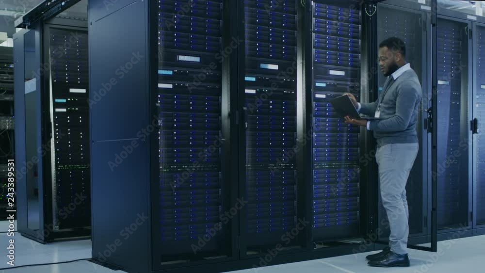 Aufkleber Black IT Specialist is Working on Laptop in Data Center while Standing Before Server Rack. Running Diagnostics, Putting in Data or Doing Maintenance Work.