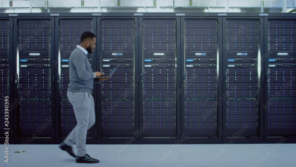 Aufkleber Black Data Center IT Technician Walking Through Server Rack Corridor with a Laptop Computer. He is Visually Inspecting Working Server Cabinets.