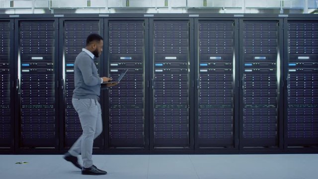 Black Data Center IT Technician Walking Through Server Rack Corridor with a Laptop Computer. He is Visually Inspecting Working Server Cabinets.