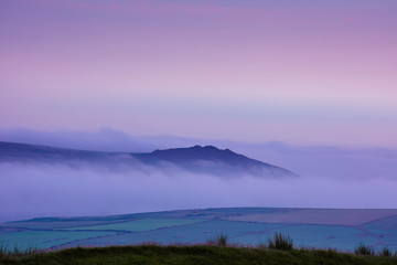 Fototapeta na wymiar Blue hour on Pembrokeshire moorlands.Hill surrounded by fog in valley and pink sky before sunrise.