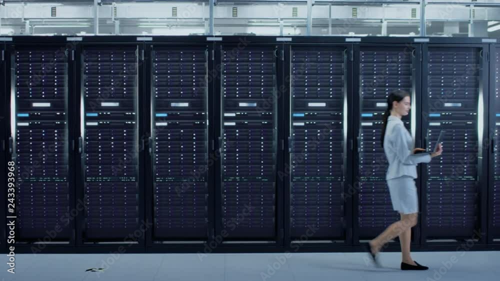 Poster female data center it specialist walking through server rack corridor with a laptop computer. she is