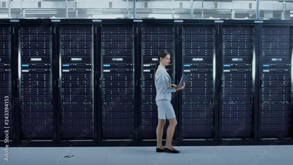 Poster Female Data Center IT Engineer Walking Through Server Rack Corridor with a Laptop Computer. She is Visually Inspecting Working Server Cabinets.