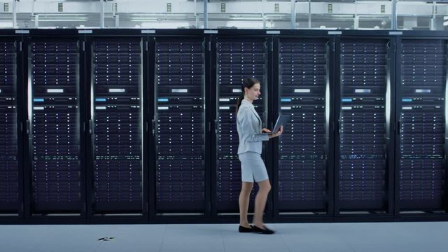 Female Data Center IT Engineer Walking Through Server Rack Corridor with a Laptop Computer. She is Visually Inspecting Working Server Cabinets.