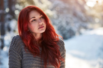 Beautiful charming girl with red hair in the winter park. Pensive, dreamy, loving look. Cold weather. Trees in the snow.