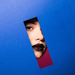 The face of a young beautiful girl with a bright make-up and puffy dark lips peers into a hole in...