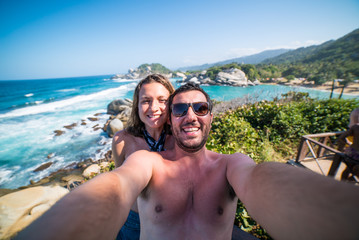 Obraz premium happy smiling couple taking selfie photo in front of the sea in Tayrona National Park, Tropical Colombia. Crazy tourists travelling on the white beach of jungles on caribbean sea. 
