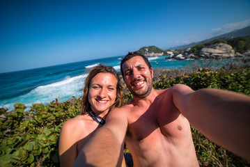 Fototapeta premium happy couple taking selfie photo in front of the sea in Tayrona National Park, Tropical Colombia. Crazy tourists travelling on the white beach of caribbean sea. 
