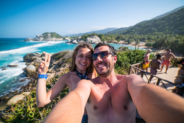 Fototapeta premium happy couple taking selfie photo in front of the sea in Tayrona National Park, Tropical Colombia. Crazy tourists travelling on the white beach of caribbean sea. 