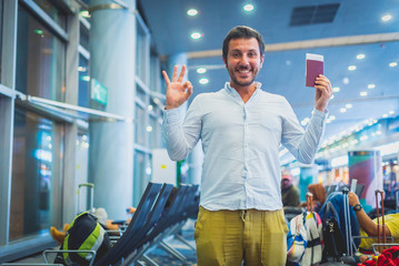 smiling man waiting in the airport, holding tickets and passport and saying OK