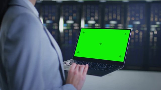 Close Up Shot of an IT Specialist Working on Laptop with Mock Up Green Screen in Data Center Next to Server Racks. Computer is Running Diagnostics or Doing Maintenance Work.