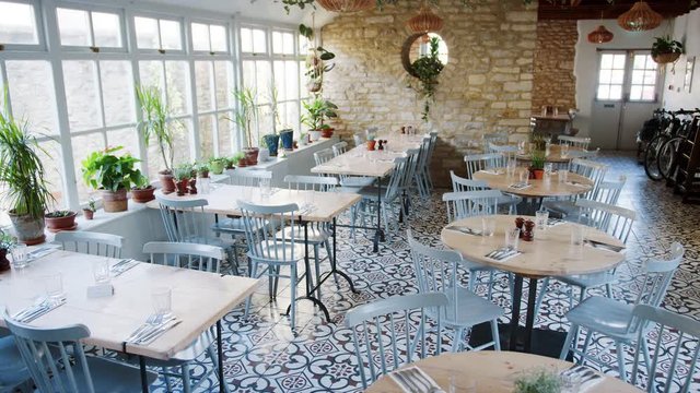 Handheld shot of empty tables and chairs by a large window in a restaurant with houseplants on the window sill, daytime 