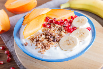 A blue plate with greek yogurt, granola, persimmon, banana, pomegranate on brown wooden background.