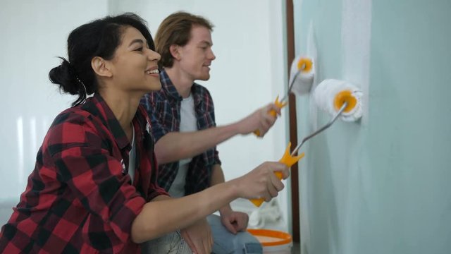 Cheerful diy couple making repair, home makeover and improvement painting interior wall with paint rollers together. Positive family stained in paint, renovating design and painting wall in new house.