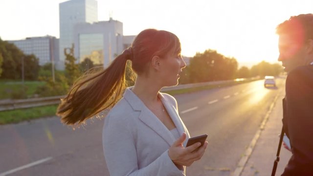 SLOW MOTION, LENS FLARE, CLOSE UP: Young woman and man crash into each other while walking down the sidewalk fixated on their smart phones. Businessman bumps into beautiful businesswoman at sunset.